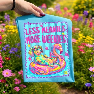 May include: A light blue t-shirt featuring a graphic of a colorful dachshund wearing heart-shaped sunglasses, floating on a pink flamingo pool float. The text reads LESS MEANIES MORE WEENIES in pink letters, surrounded by pink stars and a wavy border.