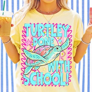 K&ouml;nnte beinhalten: Ein hellgelbes T-Shirt mit einer Grafik einer Meeresschildkr&ouml;te mit buntem Panzer. Der Text "TURTLEY DONE WITH SCHOOL!" steht in gro&szlig;en hellblauen Buchstaben darauf. Das Design enth&auml;lt rosa Wellenr&auml;nder, kleine Sterne und goldene Glitzerakzente.