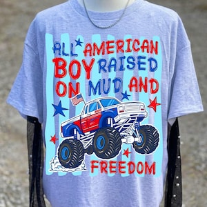 May include: A light grey short-sleeve t-shirt featuring a graphic of a red, white, and blue monster truck with a small American flag. The text reads ALL AMERICAN BOY RAISED ON MUD AND FREEDOM, surrounded by blue and red stars and vertical stripes.