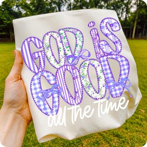 K&ouml;nnte beinhalten: Cremefarbenes Sweatshirt mit dem Schriftzug "GOD IS GOOD all The Time" in einem Patchwork-Design. Die Buchstaben weisen Blumen-, Streifen- und Vichy-Muster in Lila und Wei&szlig; auf, akzentuiert mit lila Schleifen.