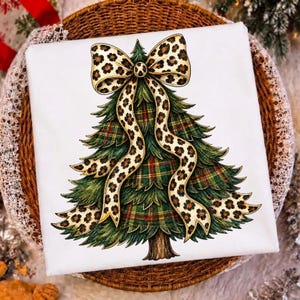 May include: A white t-shirt featuring a graphic illustration of a green Christmas tree with a red and green plaid pattern. The tree is adorned with a large, flowing bow featuring a tan and black leopard print pattern draped over the branches.