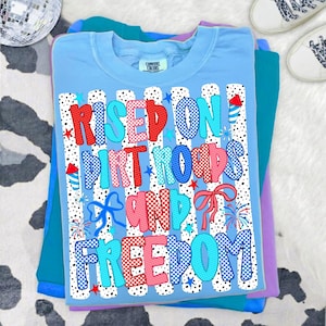May include: A light blue t-shirt featuring a graphic print with the text "RISED ON DIRT ROADS AND FREEDOM". The design includes red, white, and blue patterns, polka-dot stripes, firework illustrations, and bows, all set against a white background.