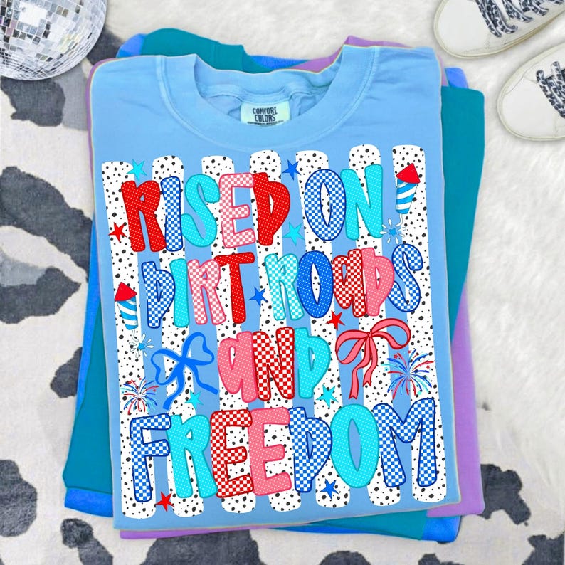 May include: A light blue t-shirt featuring a graphic print with the text "RISED ON DIRT ROADS AND FREEDOM". The design includes red, white, and blue patterns, polka-dot stripes, firework illustrations, and bows, all set against a white background.