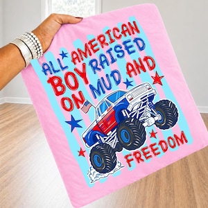 May include: A light pink blanket featuring a graphic of a red, white, and blue monster truck with large tires. The text reads ALL AMERICAN BOY RAISED ON MUD AND FREEDOM, accompanied by blue and red stars and a small American flag on the truck.