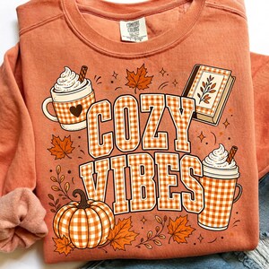 Puede incluir: Una sudadera de cuello redondo color melocot&oacute;n con un estampado gr&aacute;fico que incluye el texto COZY VIBES en un patr&oacute;n de cuadros vichy blanco y naranja. El dise&ntilde;o muestra dos tazas con nata montada, una calabaza, un libro y hojas de oto&ntilde;o.
