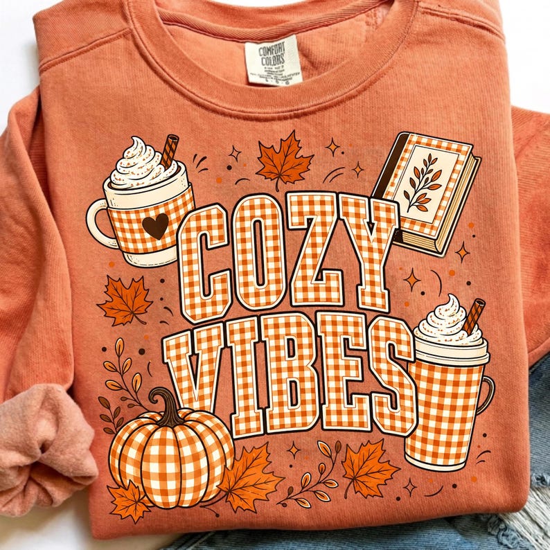 Puede incluir: Una sudadera de cuello redondo color melocot&oacute;n con un estampado gr&aacute;fico que incluye el texto COZY VIBES en un patr&oacute;n de cuadros vichy blanco y naranja. El dise&ntilde;o muestra dos tazas con nata montada, una calabaza, un libro y hojas de oto&ntilde;o.