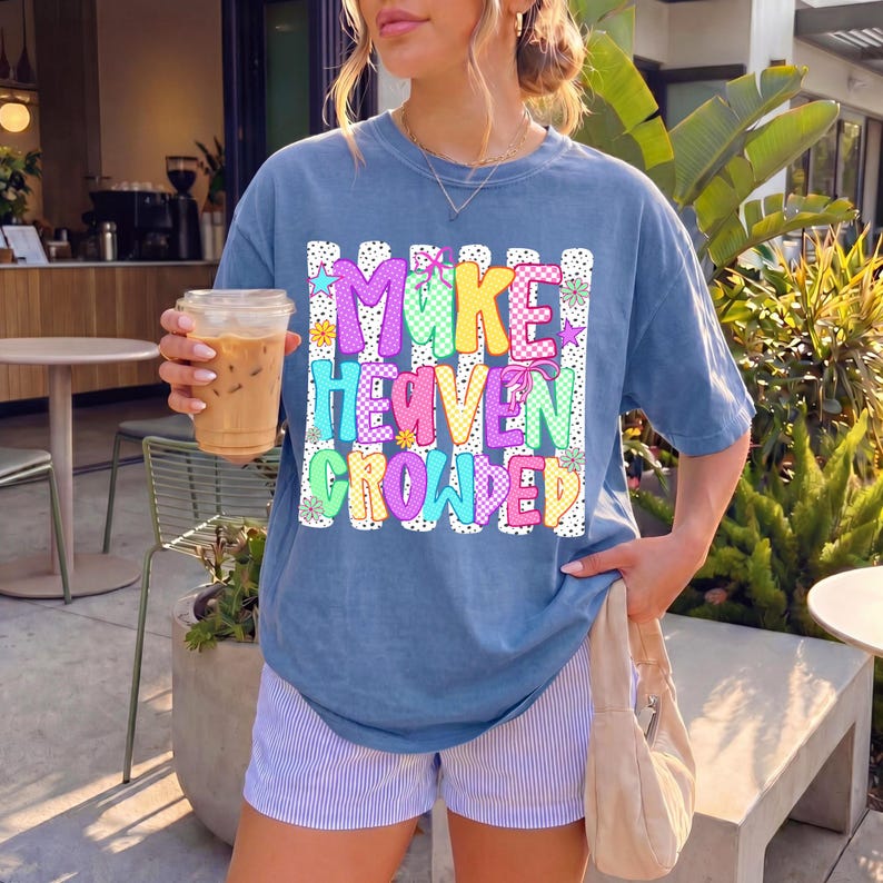 May include: A blue short-sleeve t-shirt featuring a graphic print with the text "MAKE HEAVEN CROWDED" in colorful, patterned letters. The design includes vertical stripes, polka dots, and small flower accents in pink, yellow, and teal.