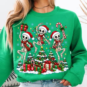 Op de afbeelding: Een felgroene sweater met ronde hals en een afbeelding van drie dansende skeletten met kerstmutsen en rood geruite sjaals. De skeletten worden omringd door kleurrijke kerstverlichting, versierde kerstbomen en ingepakte cadeaus.