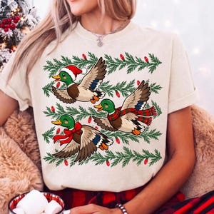 May include: A cream-colored t-shirt featuring a graphic of three mallard ducks in flight. The ducks are wearing festive holiday accessories including a Santa hat, a red and green plaid scarf, and a red bow. Green pine branches and red lights surround them.