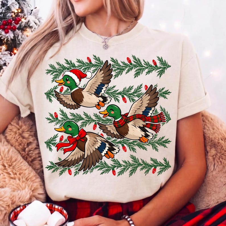 May include: A cream-colored t-shirt featuring a graphic of three mallard ducks in flight. The ducks are wearing festive holiday accessories including a Santa hat, a red and green plaid scarf, and a red bow. Green pine branches and red lights surround them.