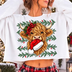 May include: A white long-sleeve sweatshirt featuring a graphic of a shaggy brown Highland cow with a red and black buffalo plaid bow on its head and a red Santa hat held in its mouth, set against a background of green pine branches and red lights.
