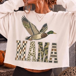 May include: A cream-colored long-sleeve sweatshirt featuring a flying mallard duck graphic above the word MAMA. The letters are filled with various patterns including a brown and white checkerboard, leopard print, camouflage, and horizontal stripes.