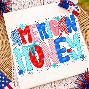 May include: A white t-shirt featuring the text AMERICAN HONEY in large, colorful, patterned letters. The design includes red, blue, and pink polka dots and checkered patterns, accented with small stars, flowers, and a firework graphic.