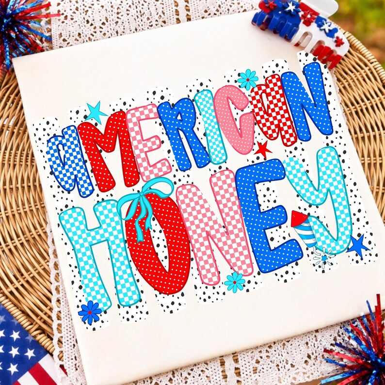May include: A white t-shirt featuring the text AMERICAN HONEY in large, colorful, patterned letters. The design includes red, blue, and pink polka dots and checkered patterns, accented with small stars, flowers, and a firework graphic.