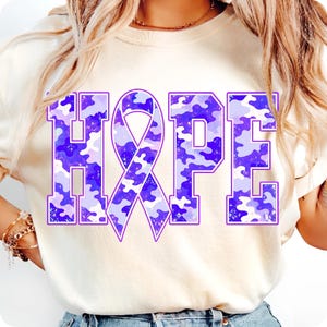 May include: A cream-coloured t-shirt featuring the word HOPE printed in large block letters. The letter O is replaced by a purple awareness ribbon. The letters and ribbon have a purple and light purple camouflage pattern with a speckled texture.