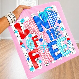 May include: A pink fabric square featuring the text LAND OF THE FREE in large, multi-colored, patterned letters. The design includes red, blue, and light blue polka dots and checkerboard patterns, accented with turquoise bows, stars, and fireworks.