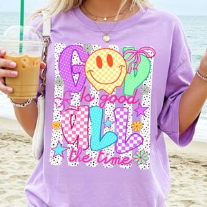 May include: A light purple t-shirt featuring a graphic print with the text God is good all the time. The design includes a yellow checkered smiley face, colorful polka-dot and checkered letters, pink bows, and small flower and star accents.