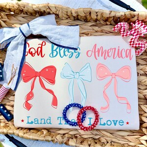 May include: A white t-shirt featuring the red and blue text God Bless America and Land That I Love, with three illustrated bows in red, light blue, and pink. The shirt is accented by a white fabric bow, a red and white checkered hair clip, and two spiral hair ties in blue and red.