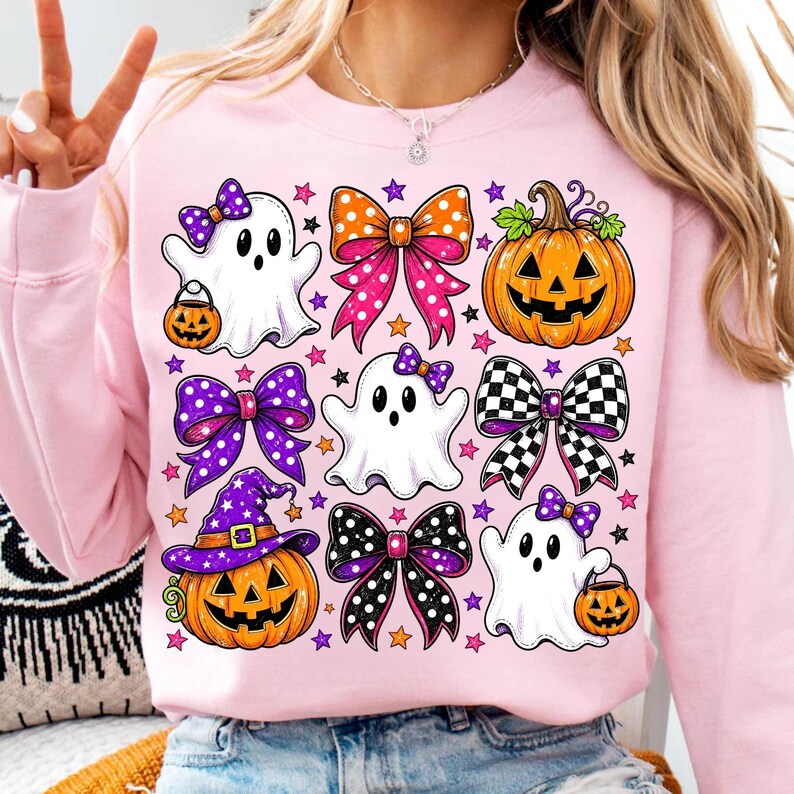 May include: A light pink sweatshirt featuring a graphic print of three white ghosts, two orange jack-o'-lanterns, and four decorative bows in various patterns including polka dots and checkered designs, all accented with small purple and orange stars.