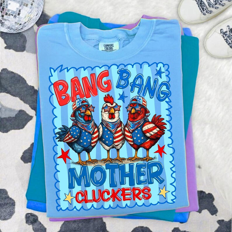 May include: A light blue t-shirt featuring a graphic of three chickens wearing American flag-themed bandanas, hats, and star-shaped sunglasses. The text reads BANG BANG MOTHER CLUCKERS in red and blue, surrounded by a blue scalloped border.
