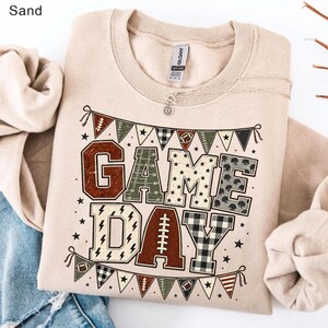 May include: A sand-colored crewneck sweatshirt featuring a graphic print of the text "GAME DAY" in large block letters. The letters are filled with various patterns including footballs, stars, and plaid. A banner of pennants hangs above and below.