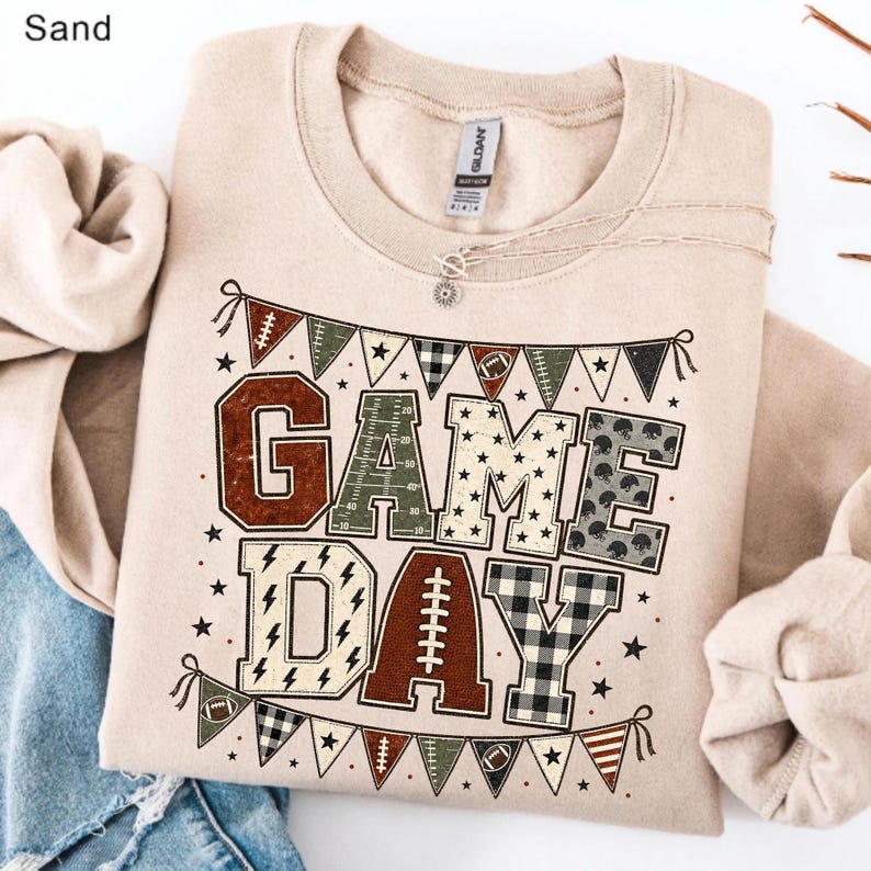 May include: A sand-colored crewneck sweatshirt featuring a graphic print of the text "GAME DAY" in large block letters. The letters are filled with various patterns including footballs, stars, and plaid. A banner of pennants hangs above and below.