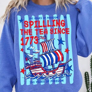 Puede incluir: Una sudadera azul de cuello redondo con un estampado gr&aacute;fico de un velero con velas rojas, blancas y azules. El texto dice 250 YEARS OF FREEDOM, SPILLING THE TEA SINCE 1773 y 250 YEARS OF FREEDOM, rodeado de estrellas rojas y azules.