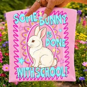 K&ouml;nnte beinhalten: Ein hellrosa Stoffartikel mit einer zentralen Illustration eines wei&szlig;en Kaninchens in einem rosa ovalen Rahmen. Der Text "SOME BUNNY IS DONE WITH SCHOOL!" ist in leuchtend blauen Buchstaben gedruckt, umgeben von rosa Wellenr&auml;ndern und gelben Sternen.