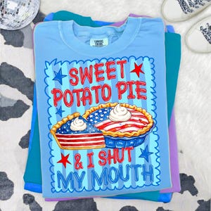 Op de afbeelding: Een lichtblauw t-shirt met een afbeelding van een hele taart en een taartpunt, beide versierd met rode en witte strepen en blauwe sterren. De tekst SWEET POTATO PIE & I SHUT MY MOUTH staat in rood en blauw, omringd door een blauwe geschulpte rand.