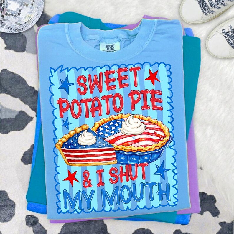 Op de afbeelding: Een lichtblauw t-shirt met een afbeelding van een hele taart en een taartpunt, beide versierd met rode en witte strepen en blauwe sterren. De tekst SWEET POTATO PIE & I SHUT MY MOUTH staat in rood en blauw, omringd door een blauwe geschulpte rand.