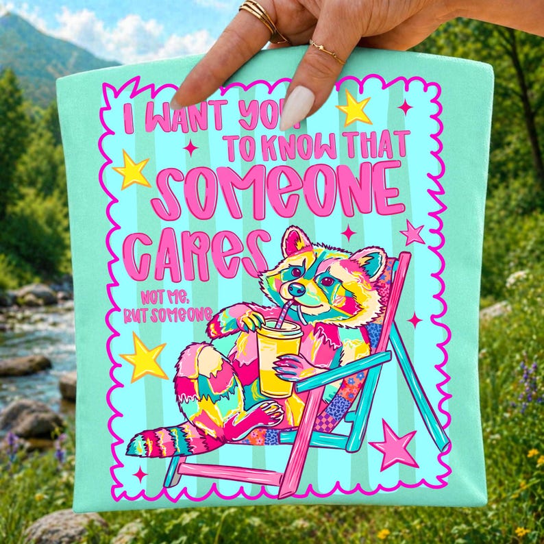 K&ouml;nnte beinhalten: Eine hellblaue Tragetasche mit einem bunten Waschb&auml;ren, der in einem Strandstuhl sitzt und ein Getr&auml;nk h&auml;lt. Der Text lautet: I WANT YOU TO KNOW THAT SOMEONE CARES NOT ME, BUT SOMEONE. Das Design enth&auml;lt rosa gewellte R&auml;nder und gelbe Sterne.