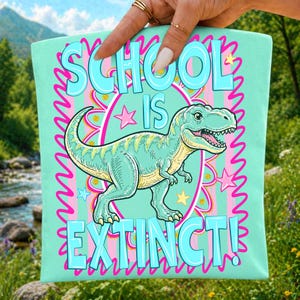 Op de afbeelding: Een licht mintgroen stoffen vierkant met een centrale illustratie van een groene Tyrannosaurus Rex. De tekst SCHOOL IS EXTINCT! is gedrukt in grote lichtblauwe letters. Het ontwerp bevat roze sterren en een golvend roze randpatroon.