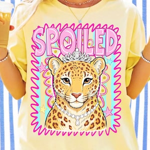 Op de afbeelding: Lichtgele t-shirt met een afbeelding van een luipaard met een kroon en een parelketting, met het woord "SPOILED" in roze letters. De luipaard staat in een roze en blauwe golvende rand.