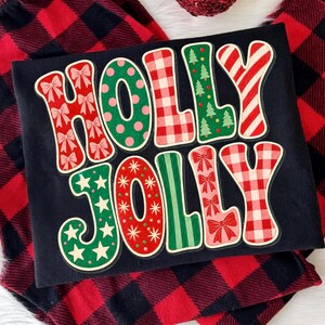 May include: A black t-shirt featuring the text HOLLY JOLLY in large, colorful, patterned letters. The letters include designs of red bows, green polka dots, red and white gingham, green pine trees, red and white stripes, stars, and red dots.