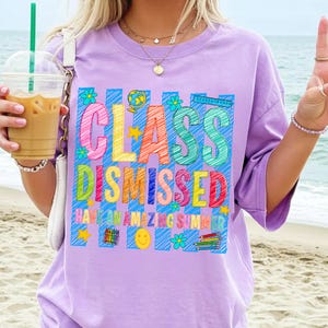 May include: A light purple t-shirt featuring a colorful graphic with the text "CLASS DISMISSED" and "HAVE AN AMAZING SUMMER". The design includes scribbled letters, a globe, a ruler, crayons, pencils, flowers, stars, and a smiley face.
