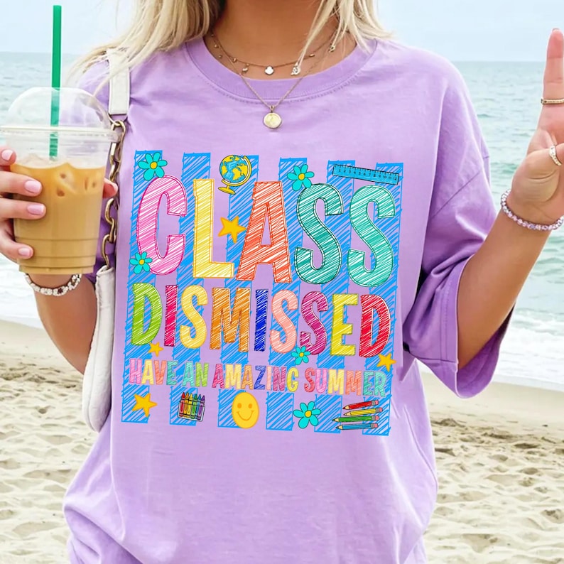 May include: A light purple t-shirt featuring a colorful graphic with the text "CLASS DISMISSED" and "HAVE AN AMAZING SUMMER". The design includes scribbled letters, a globe, a ruler, crayons, pencils, flowers, stars, and a smiley face.