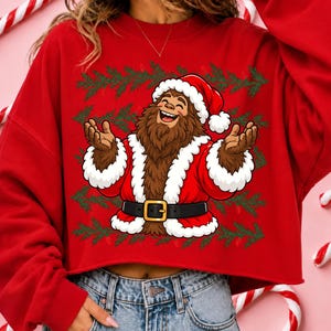 Puede incluir: Una sudadera roja de manga larga con un gr&aacute;fico de un personaje Bigfoot marr&oacute;n vestido con un traje de Santa Claus, que incluye un gorro rojo con borde blanco, un cintur&oacute;n negro con hebilla dorada y pu&ntilde;os de piel blanca sobre ramas de pino verdes.