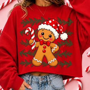Puede incluir: Una sudadera roja de manga larga con el gr&aacute;fico de un hombre de jengibre que lleva un gorro de Pap&aacute; Noel rojo con lunares blancos y una pajarita roja. El hombre de jengibre sostiene un bast&oacute;n de caramelo rojo y blanco sobre ramas de pino.