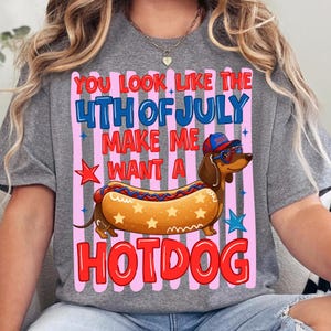 Op de afbeelding: Een grijs gem&ecirc;leerd t-shirt met een afbeelding van een teckel met een baseballpet en hartvormige zonnebril, met een hotdog-lichaam. De tekst luidt: YOU LOOK LIKE THE 4TH OF JULY MAKE ME WANT A HOTDOG, omringd door roze strepen en sterren.