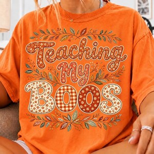 Pu&ograve; includere: Una t-shirt arancione a maniche corte con il testo "Teaching My BOOS" in un grande carattere decorativo. Le lettere sono riempite con motivi floreali e a quadri nei toni dell'arancione, marrone e bianco, circondate da foglie autunnali.
