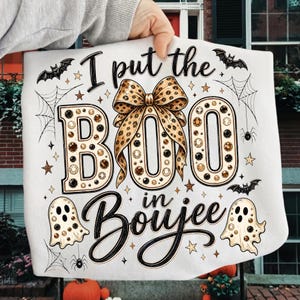 Pu&ograve; includere: Una t-shirt bianca con una grafica che riporta il testo I put the BOO in Boujee. Il design include un grande fiocco con stampa leopardata, due fantasmi con macchie leopardate, pipistrelli neri, ragnatele, ragni e piccole stelle dorate.