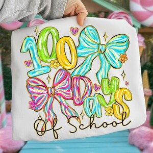 Op de afbeelding: Wit t-shirt met de tekst "100 Days of School" in kleurrijke letters en decoratieve strikken. Het ontwerp bevat groene, gele, blauwe en roze elementen, met gouden accenten en kleine hartjes.