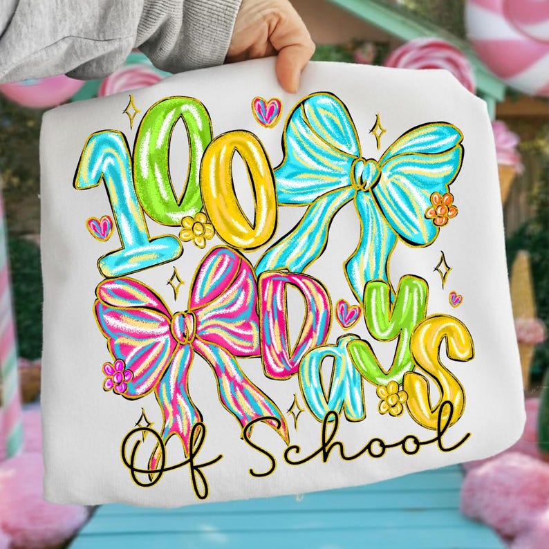 Op de afbeelding: Wit t-shirt met de tekst "100 Days of School" in kleurrijke letters en decoratieve strikken. Het ontwerp bevat groene, gele, blauwe en roze elementen, met gouden accenten en kleine hartjes.