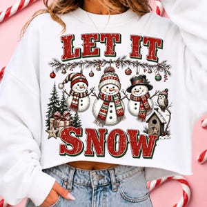 Pu&ograve; includere: Una felpa bianca a maniche lunghe con una stampa grafica che riporta il testo LET IT SNOW in rosso e verde. Il design include tre pupazzi di neve con cappelli e sciarpe, decorazioni appese, un pino, un regalo, una casetta e un gufo.