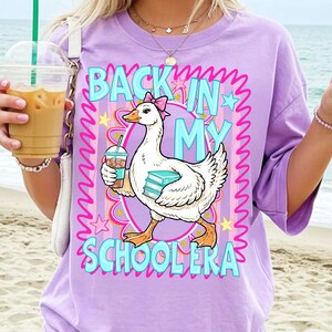 May include: A lilac-colored t-shirt with the phrase "BACK IN MY SCHOOLERA" in turquoise and pink. A cartoon goose wearing a pink bow holds a coffee and books. The shirt has short sleeves and a crew neck.