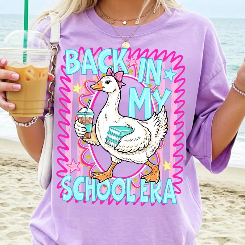 May include: A lilac-colored t-shirt with the phrase "BACK IN MY SCHOOLERA" in turquoise and pink. A cartoon goose wearing a pink bow holds a coffee and books. The shirt has short sleeves and a crew neck.