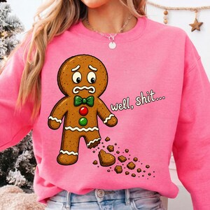 May include: A bright pink crewneck sweatshirt featuring a cartoon gingerbread person with a worried expression, a green bow tie, and red and green buttons. The gingerbread person has a missing foot with crumbs, and the text reads well, shit...