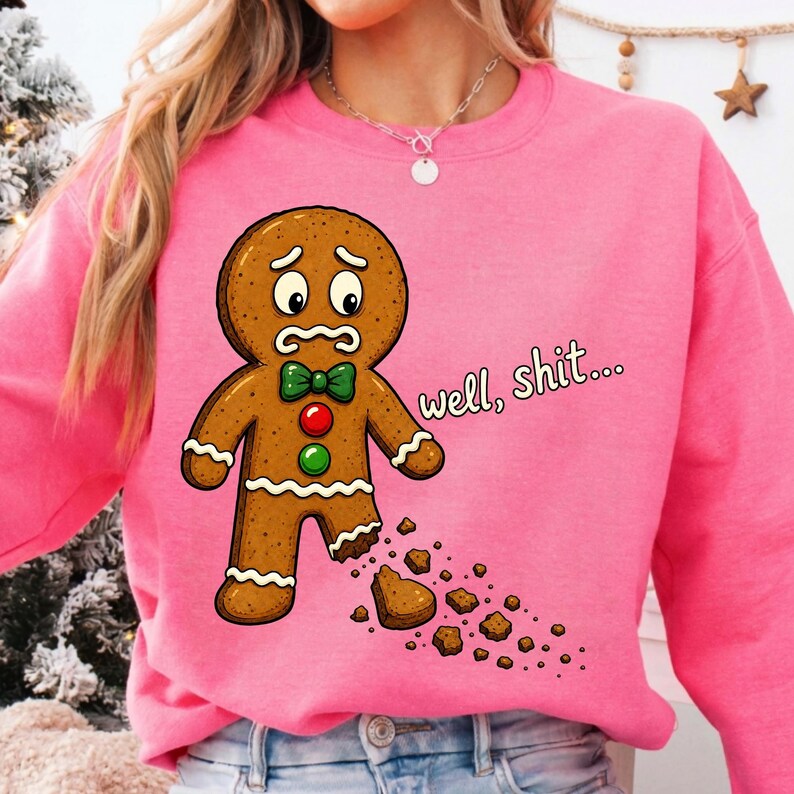 May include: A bright pink crewneck sweatshirt featuring a cartoon gingerbread person with a worried expression, a green bow tie, and red and green buttons. The gingerbread person has a missing foot with crumbs, and the text reads well, shit...
