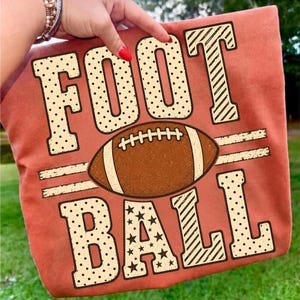 May include: A terracotta-colored t-shirt featuring the text FOOTBALL in large, decorative block letters. The letters have varied patterns including polka dots, diagonal stripes, and stars. A brown football graphic sits between the two words.