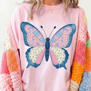 May include: A light pink t-shirt featuring a large butterfly design. The butterfly has blue denim-like edges, and the wings are filled with floral and plaid patterns in pink, purple, and blue. The sleeves of a colorful sweater are visible.